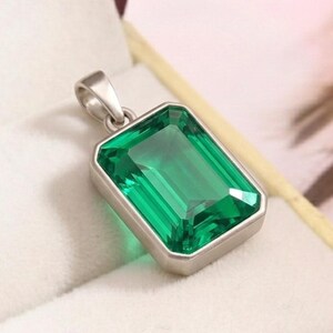 May include: A rectangular, emerald-green gemstone pendant with a silver setting. The pendant is presented in a white jewellery box with a gold trim. The gemstone has a faceted surface that reflects light.