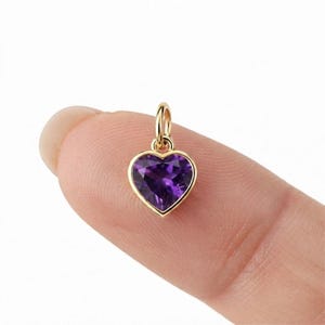 May include: A small heart-shaped pendant featuring a faceted deep purple amethyst gemstone set in a polished yellow gold frame with a matching gold jump ring attached to the top, designed for use as a charm on a necklace or bracelet chain.