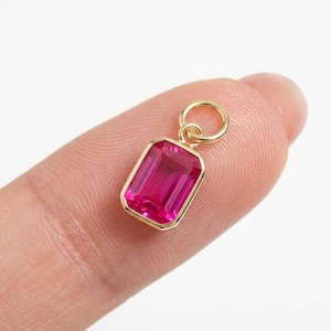 May include: A rectangular emerald-cut pendant featuring a vibrant pink gemstone set in a polished gold-toned metal frame. The pendant includes a small circular jump ring attached to the top, allowing it to be easily suspended from a chain or cord.