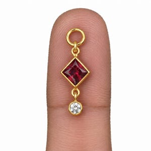 Könnte beinhalten: Ein Goldanhänger mit einem quadratisch geschliffenen, tiefroten Edelstein in einer Goldfassung, ergänzt durch einen kleinen, runden, klaren Edelstein, der unten hängt, sowie einem goldenen Verbindungsring an der Oberseite.
