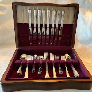 May include: A wooden silverware case with a burgundy velvet interior. The case is open, revealing a set of silver-colored utensils, including knives, forks, and spoons. The silverware has decorative handles. The case is closed with a wooden lid.