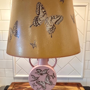 May include: A decorative table lamp with a pink base and a tan lampshade. The lampshade and base are adorned with butterfly illustrations in black and white. The lamp has a classic, vintage design.