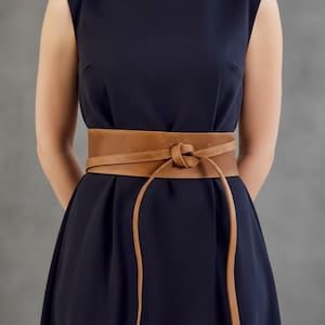 Op de afbeelding: Een lichtbruine leren wikkelriem, vooraan geknoopt, gedragen over een marineblauwe jurk. De lange riemen van de riem hangen naar beneden en creëren een stijlvol accessoire. De riem is gemaakt van zacht leer.