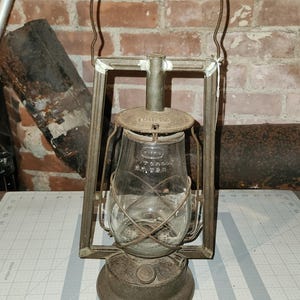 May include: A vintage metal Dietz lantern with a clear glass globe, a wire handle, and a circular base. The glass globe features embossed text reading Dietz and N.Y. U.S.A. The lantern has a weathered, dark grey finish and a sturdy frame structure.