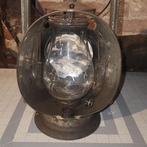 May include: A weathered metal railroad lantern featuring a clear glass globe, a circular reflector, and a rectangular top handle. The lantern is positioned on a cutting mat with visible markings, showing a width of approximately 8 inches.