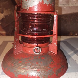 May include: A vintage red metal lantern featuring a cylindrical red glass globe with the embossed text "Dietz No 50". The lantern has a wide, circular base and a protective metal cage surrounding the glass, showing signs of weathered paint.