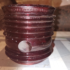 May include: A deep red glass Fresnel lens for a nautical light, featuring a cylindrical shape with horizontal ridges and a circular indentation on the side. The top rim is embossed with the text "NIGHT WATCH FRESNEL" in clear, raised lettering.