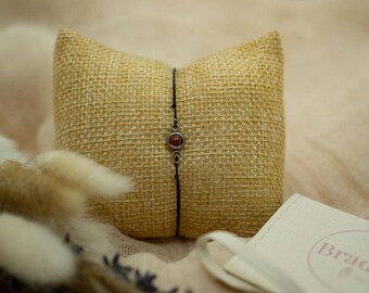 Puede incluir: Una delicada pulsera con una pequeña gema redonda de color marrón rojizo engastada en un metal plateado. La pulsera se exhibe sobre una pequeña almohada cuadrada de arpillera beige. Flores secas y una pequeña bolsa con el texto "Braddict" también están en la imagen.
