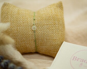 May include: A light green bracelet with a circular pendant and silver accents is displayed on a small, burlap pillow. The pillow is set against a soft, neutral background, with a small card featuring the brand name "Braddict".
