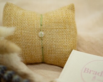 May include: A delicate bracelet with a light green cord and a round, pale gemstone bead is displayed on a small, textured, tan pillow. The bracelet is centered on the pillow, with a small, off-white box with the word "Braddict" visible.