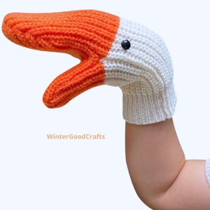 Op de afbeelding: Een gebreide ganzenpop met een oranje snavel en een witte kop en nek. De pop heeft een zwart knoopoog en is ontworpen om over een hand te passen. De tekst "WinterGoodCrafts" staat onderaan.