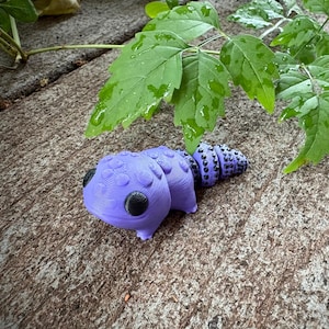 May include: A purple caterpillar toy with black eyes and a segmented body. The toy is resting on a textured, brown surface, with green leaves in the background.