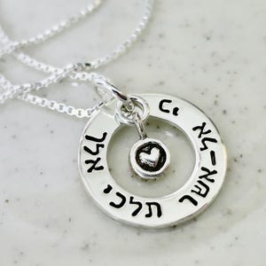 May include: A silver necklace with a circular pendant and a small heart charm. The pendant has Hebrew text around the edge and a heart-shaped charm in the center. The necklace is displayed on a white, textured surface.