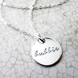 May include: A silver necklace with a round pendant that says "bubbie" in cursive.