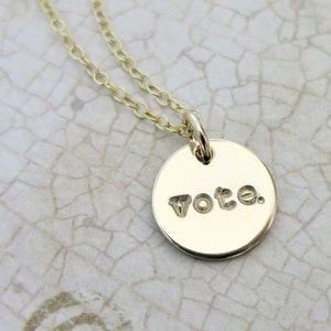 A small round gold pendant necklace sits on an aged cream paper. On the pendant is the word "vote" with a period after it in a lowercase typewriter font. Attached to the pendant is a delicate gold cable chain.