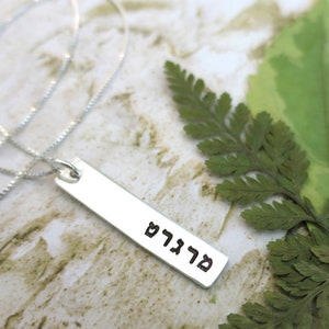 Op de afbeelding: Een zilveren ketting met een rechthoekige hanger waarop "שלום" in het Hebreeuws staat.