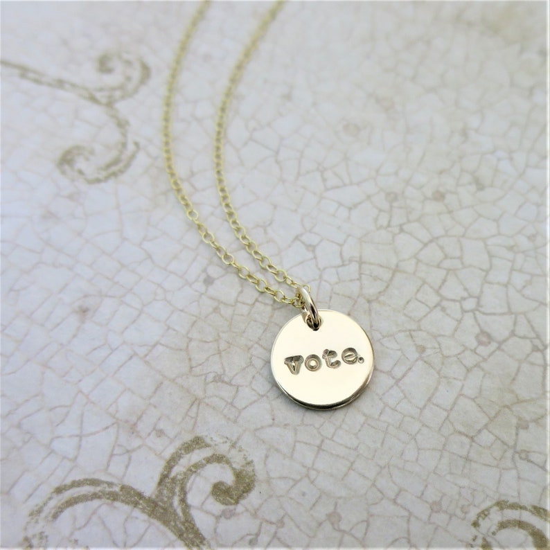 A small round gold pendant necklace sits on an aged cream paper. On the pendant is the word "vote" with a period after it in a lowercase typewriter font. Attached to the pendant is a delicate gold cable chain.