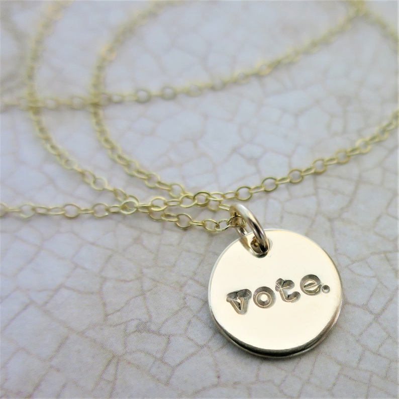 A small round gold pendant necklace sits on an aged cream paper. On the pendant is the word "vote" with a period after it in a lowercase typewriter font. Attached to the pendant is a delicate gold cable chain.