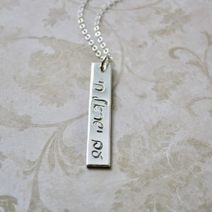 May include: A silver necklace with a rectangular pendant inscribed with Hebrew text. The pendant is hanging on a silver chain.
