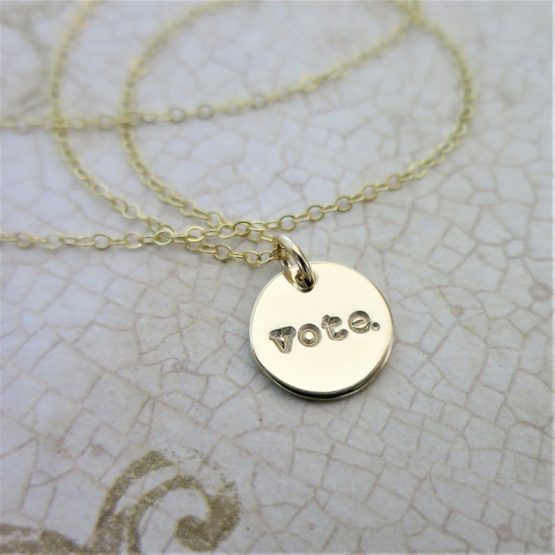 A small round gold pendant necklace sits on an aged cream paper. On the pendant is the word "vote" with a period after it in a lowercase typewriter font. Attached to the pendant is a delicate gold cable chain.