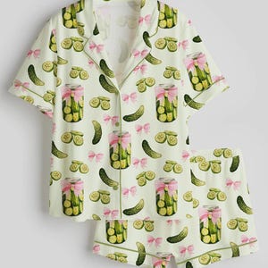 May include: A two-piece pajama set featuring a light green background with a pattern of pickles, pickle slices, jars of pickles tied with pink bows. The set includes a short-sleeved button-up shirt and matching shorts.