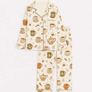 May include: A two-piece pajama set in cream fabric featuring a repeating pattern of brown and green teapots, teacups, and tea bags. The long-sleeved button-down shirt has a collar and a chest pocket, while the matching pants have a straight-leg cut.
