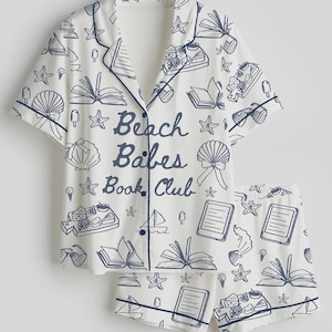 May include: White pajama set with a navy blue trim and a repeating pattern of books, seashells, ice cream, and sailboats. The shirt reads "Beach Babes Book Club" in navy blue script.