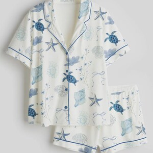 May include: White pyjama set with a blue sea life pattern. The short-sleeved top and shorts feature blue turtles, seashells, and starfish. The set has blue trim and buttons.