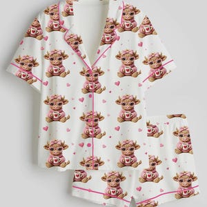 May include: White pajama set with a repeating pattern of cartoon cows wearing pink sweaters and holding coffee cups. The set includes a short-sleeved top and shorts, both trimmed with pink piping and scattered pink hearts.