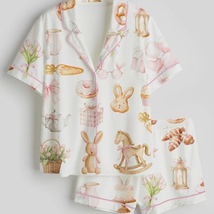 May include: White pajama set with a whimsical Easter-themed print. The short-sleeved top and shorts feature pink trim and are covered in illustrations of bunnies, carrots, and gifts. The set is designed for comfort and a festive feel.
