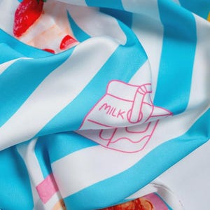 Puede incluir: Pa&ntilde;uelo de seda con un patr&oacute;n repetido de rayas azules y blancas, fresas y una ilustraci&oacute;n de cart&oacute;n de leche. El pa&ntilde;uelo tiene una textura suave y fluida y una paleta de colores vibrantes.
