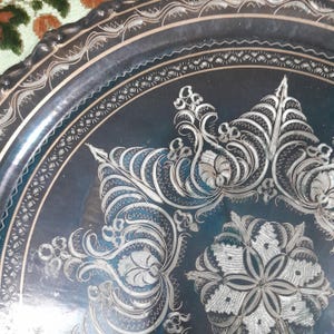 May include: Close-up of a decorative, dark blue metal tray with intricate silver floral and geometric patterns. The tray's edge has a scalloped design. The detailed carvings and contrasting colors create a vintage aesthetic.