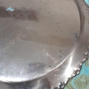 May include: A tarnished silver-colored metal tray with a scalloped edge. The tray has a circular shape with visible scratches and imperfections on its surface. The edge features a decorative floral design.