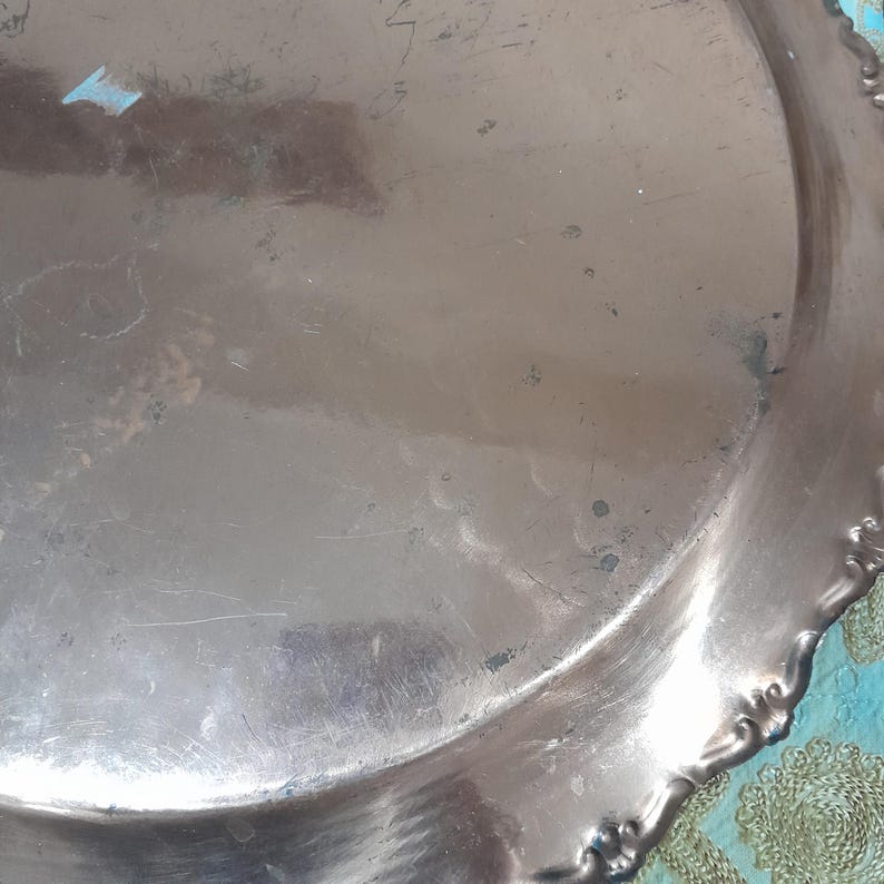 May include: A tarnished silver-colored metal tray with a scalloped edge. The tray has a circular shape with visible scratches and imperfections on its surface. The edge features a decorative floral design.