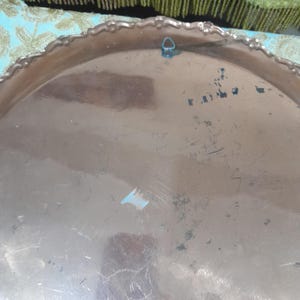 May include: A tarnished, round metal tray with a scalloped edge. The tray has a silver finish with visible scratches and wear. A small blue loop is attached to the top edge for hanging. The tray's surface reflects light, showing its age and use.