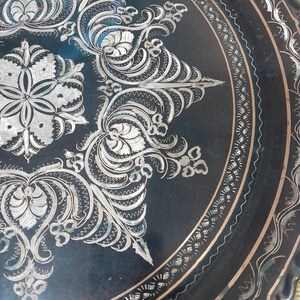May include: Close-up of a black metal tray with intricate silver floral and scrollwork designs. The tray features a detailed border with a repeating pattern. The design is symmetrical and ornate, with a vintage aesthetic.