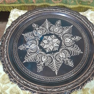 May include: A dark metal tray with intricate silver floral designs. The tray has a scalloped edge and a central starburst pattern. The detailed engraving adds a decorative touch, suitable for serving or display.