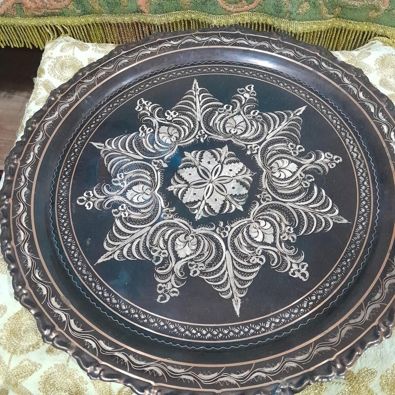 May include: A dark metal tray with intricate silver floral designs. The tray has a scalloped edge and a central starburst pattern. The detailed engraving adds a decorative touch, suitable for serving or display.