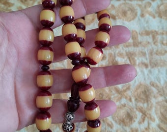 Pode incluir: Um fio de contas de oração com contas ovais amarelas e marrons alternadas. As contas são enfiadas juntas, com um pingente decorativo prateado e uma peça de madeira marrom escura na extremidade.