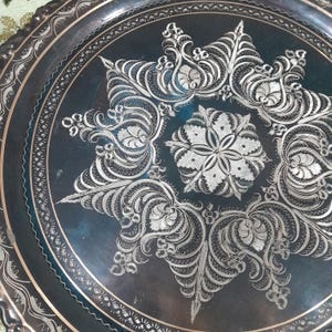 May include: A detailed close-up of a decorative metal tray. The tray is dark with intricate silver floral and geometric patterns. The design is centered with a starburst pattern and surrounded by a detailed border.