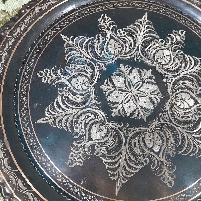 May include: A detailed close-up of a decorative metal tray. The tray is dark with intricate silver floral and geometric patterns. The design is centered with a starburst pattern and surrounded by a detailed border.