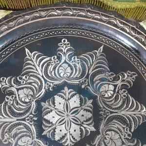 May include: A dark, round metal tray with intricate silver floral and scrollwork designs. The tray's edge features a decorative border. The detailed patterns and contrasting colors create a visually appealing piece, suitable for display or serving.