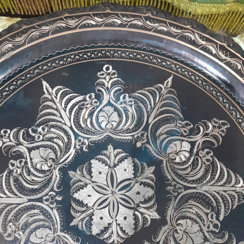 May include: A dark, round metal tray with intricate silver floral and scrollwork designs. The tray's edge features a decorative border. The detailed patterns and contrasting colors create a visually appealing piece, suitable for display or serving.