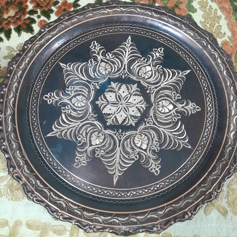May include: A round, silver-toned metal tray with an intricate, etched floral design. The tray features a dark center with a detailed, symmetrical pattern and a scalloped edge. The decorative tray is likely used for serving or display.