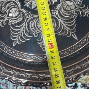 May include: A decorative, dark metal plate with intricate silver floral and geometric patterns. The plate's edge has a scalloped design. A yellow ruler shows a measurement of approximately 12 inches.
