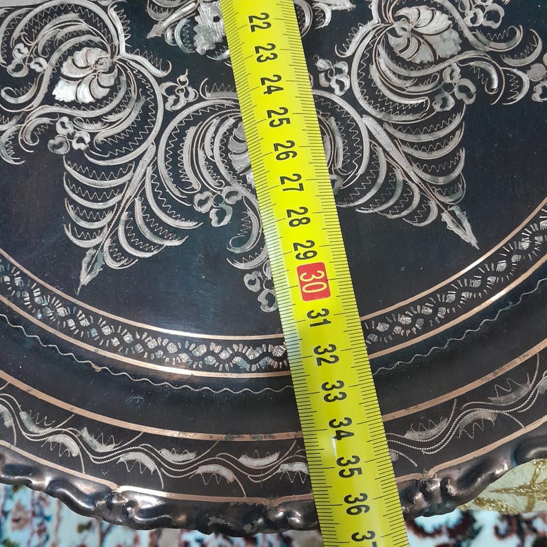 May include: A decorative, dark metal plate with intricate silver floral and geometric patterns. The plate's edge has a scalloped design. A yellow ruler shows a measurement of approximately 12 inches.