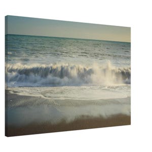 May include: A canvas print showing a coastal scene. A wave is breaking on a sandy shore. The water is a blend of turquoise and white, with a pale blue sky. The sand is a dark brown colour.