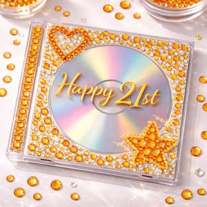 May include: A clear CD case decorated with orange and clear rhinestones. The CD has the words "Happy 21st" in gold script. A heart and star are also made of orange rhinestones.