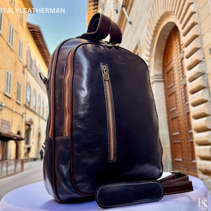 Puede incluir: Una mochila de cuero azul marino con detalles y cremalleras marrones. La mochila tiene un asa superior y una correa para el hombro desmontable. El texto "ITALYLEATHERMAN" es visible en la parte superior izquierda.