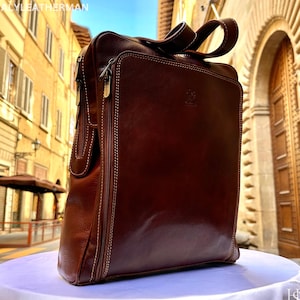 May include: A brown leather messenger bag with a top handle and a front zip pocket. The bag features white stitching and a logo. The bag is on a white surface. The text "ITALY LEATHERMAN" is visible at the top.